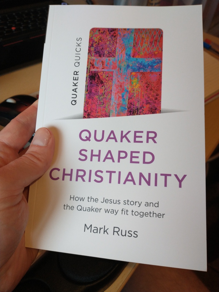 My hand holding a copy of my book, Quaker Shaped Christianity, with a white cover, purple text and a pink and blue image of a cross.